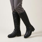 A pair of black knee-high leather boots with side zippers and a buckle strap detail on the lower leg, shown on a model wearing gray trousers against a light beige studio background.