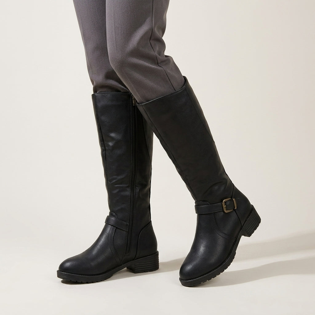 A pair of black knee-high leather boots with side zippers and a buckle strap detail on the lower leg, shown on a model wearing gray trousers against a light beige studio background.