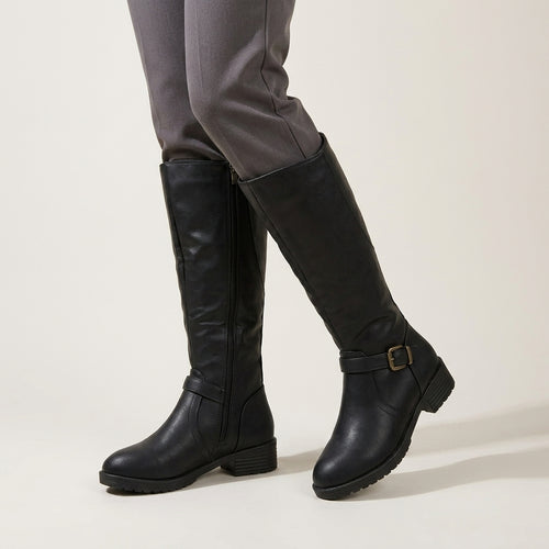 A pair of black knee-high leather boots with side zippers and a buckle strap detail on the lower leg, shown on a model wearing gray trousers against a light beige studio background.