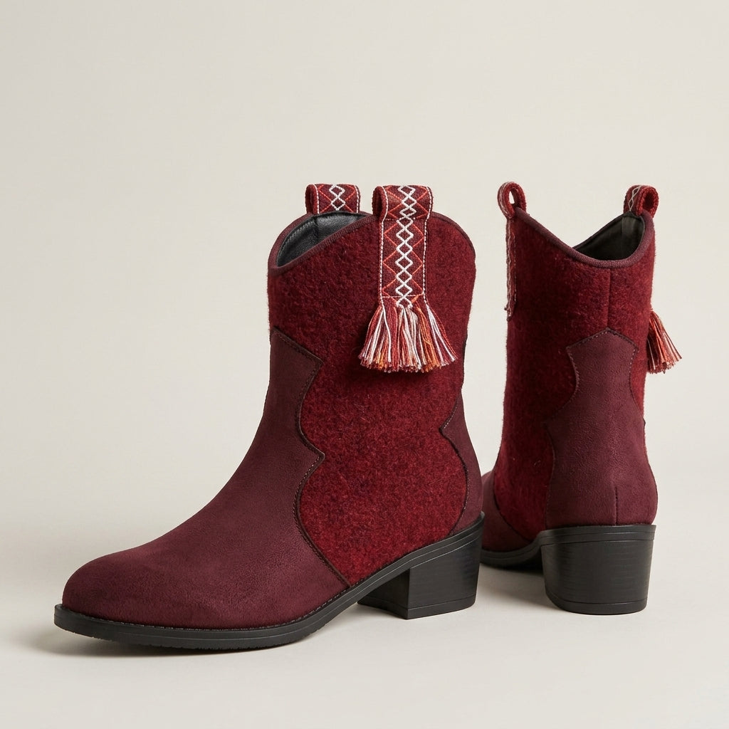 A pair of women's western-style ankle boots in deep burgundy, featuring decorative woven trim and tassels on the pull tabs, set against a plain light beige background.