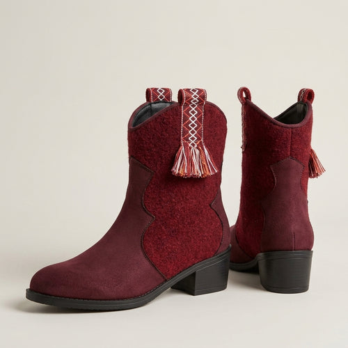 A pair of women's western-style ankle boots in deep burgundy, featuring decorative woven trim and tassels on the pull tabs, set against a plain light beige background.