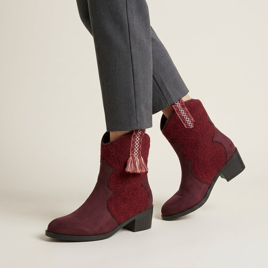 A pair of women's western-style ankle boots in burgundy, shown on a model wearing gray trousers against a light beige studio background.