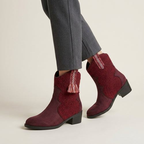 A pair of women's western-style ankle boots in burgundy, shown on a model wearing gray trousers against a light beige studio background.