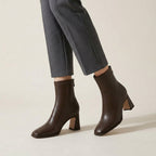 A person wearing dark brown leather ankle boots with block heels and gray cropped trousers, shown from the calves down against a plain light beige studio background.