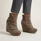 A pair of taupe-brown suede wedge ankle boots with buckle straps, shown on a model wearing grey trousers against a light beige studio background.