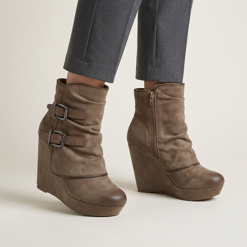 A pair of taupe-brown suede wedge ankle boots with buckle straps, shown on a model wearing grey trousers against a light beige studio background.